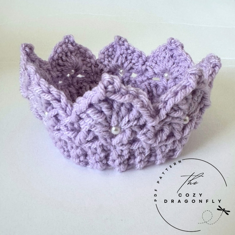 May include: A lavender crocheted crown with a scalloped edge and pearl embellishments. The crown is made with a delicate stitch pattern and has a soft, feminine look.
