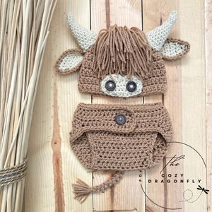 May include: A crocheted brown and white highland cow hat and diaper set for a baby. The hat has a brown fuzzy mane and a white face with black button eyes. The diaper has a brown crocheted tail.