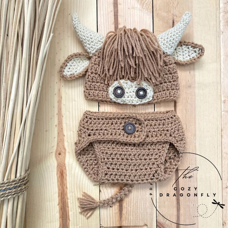 May include: A crocheted brown and white highland cow hat and diaper set for a baby. The hat has a brown fuzzy mane and a white face with black button eyes. The diaper has a brown crocheted tail.