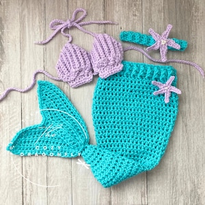May include: A crocheted mermaid costume set for a baby. The set includes a purple shell-shaped bra top, a turquoise mermaid tail, and a headband with a starfish.