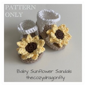 K&ouml;nnte beinhalten: Ein Paar geh&auml;kelte Babysandalen mit Sonnenblumenverzierungen. Die Sandalen sind wei&szlig; und beige mit gelben und braunen geh&auml;kelten Sonnenblumen.