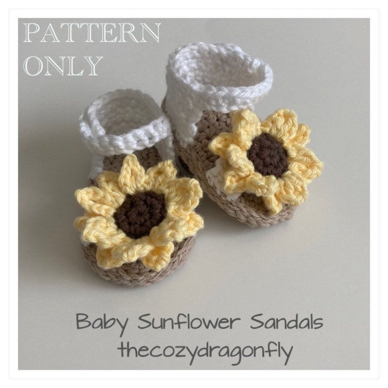 K&ouml;nnte beinhalten: Ein Paar geh&auml;kelte Babysandalen mit Sonnenblumenverzierungen. Die Sandalen sind wei&szlig; und beige mit gelben und braunen geh&auml;kelten Sonnenblumen.