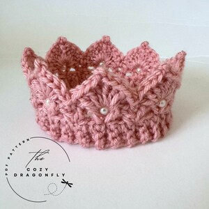 May include: A pink crocheted crown with white pearl embellishments. The crown has a scalloped edge and a delicate, intricate design. The crown is handmade and would be a beautiful accessory for a special occasion.