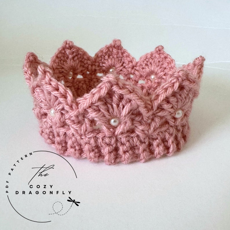 May include: A pink crocheted crown with white pearl embellishments. The crown has a scalloped edge and a delicate, intricate design. The crown is handmade and would be a beautiful accessory for a special occasion.