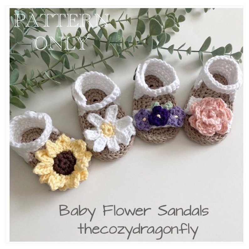 K&ouml;nnte beinhalten: Vier geh&auml;kelte Babysandalen mit Blumenverzierungen. Die Sandalen sind in den Farben Wei&szlig;, Braun, Gelb, Rosa, Lila und Gr&uuml;n gehalten. Die Sandalen sind in einer Reihe auf einer wei&szlig;en Oberfl&auml;che angeordnet. Der Text "Baby Flower Sandals" und "thecozydragonfly" befindet sich unter den Sandalen.