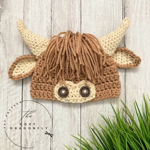 May include: A crocheted brown and cream highland cow hat with brown button eyes. The hat is on a white wood background with green grass in the foreground.