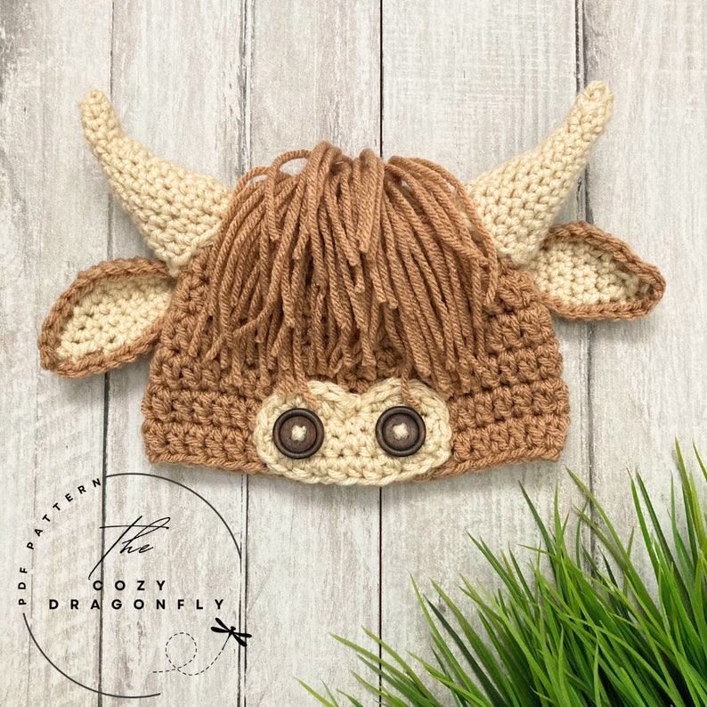 May include: A crocheted brown and cream highland cow hat with brown button eyes. The hat is on a white wood background with green grass in the foreground.