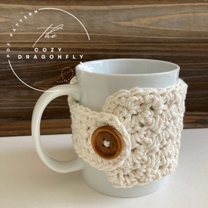 May include: A white ceramic mug with a crocheted beige cozy featuring a wooden button. The cozy is crocheted in a square pattern.