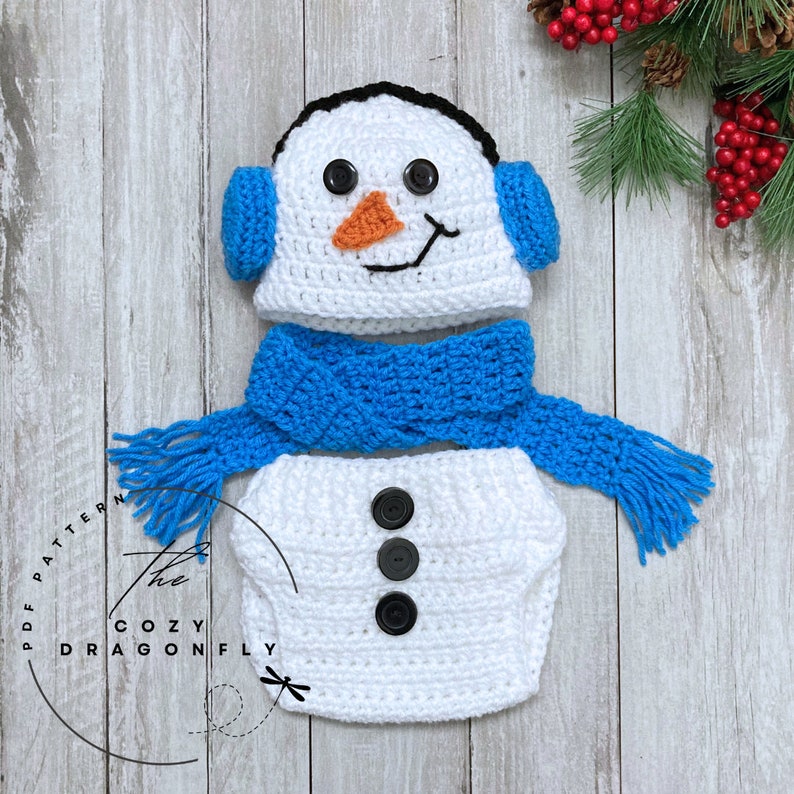 May include: A white crocheted snowman outfit for a baby, featuring a hat with blue earmuffs, a blue scarf, and a white diaper cover with black buttons.