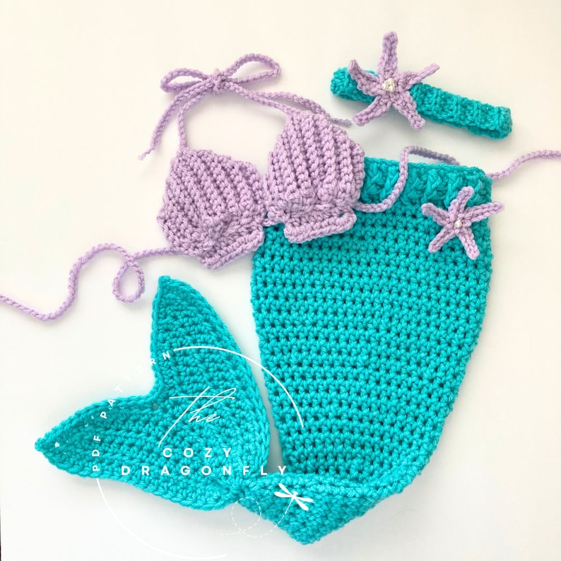 May include: A crocheted mermaid costume for a baby, featuring a purple top, a turquoise tail, and a headband with a starfish.