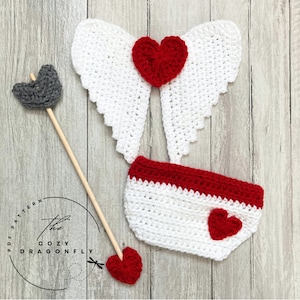 May include: A crochet baby outfit with white wings, a red heart, and a red and white diaper cover. The outfit is accompanied by a wooden arrow with a gray felt arrowhead and a red felt heart.