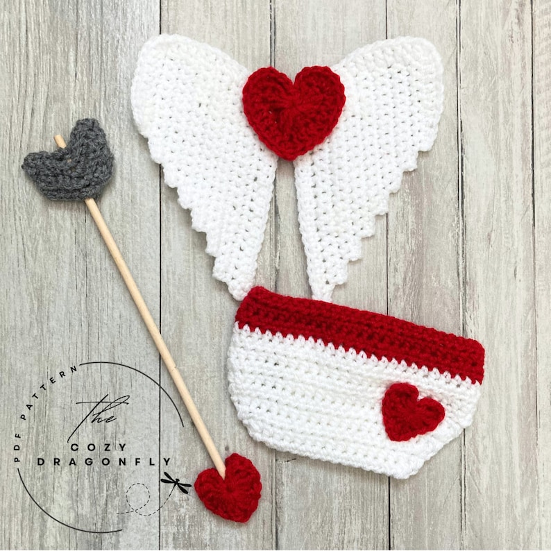 May include: A crochet baby outfit with white wings, a red heart, and a red and white diaper cover. The outfit is accompanied by a wooden arrow with a gray felt arrowhead and a red felt heart.