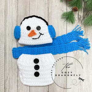 May include: A white crocheted snowman outfit for a baby, featuring a hat with ear flaps, a scarf, and three black buttons. The hat has a black outline and an orange nose. The scarf is blue with white fringe. The outfit is on a wooden background.