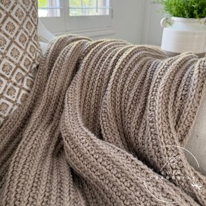 May include: A cozy, ribbed knit blanket in a neutral taupe color. The blanket is draped over a sofa, showcasing its thick, textured weave. The corner of a patterned pillow is visible, along with a glimpse of a window and a plant in a white pot.