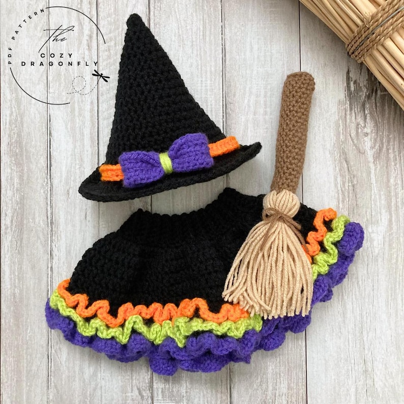 May include: Crocheted Halloween witch costume set, including a black pointed hat with purple bow and orange trim, a black skirt with purple, green, and orange trim, and a brown broom. The Cozy Dragonfly pattern is visible.
