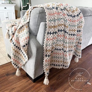Puede incluir: Una manta de ganchillo color crema con un patrón geométrico en azul marino, rosa, dorado y turquesa. La manta está sobre un sofá gris y tiene borlas en las esquinas. El logotipo de Cozy Dragonfly es visible.