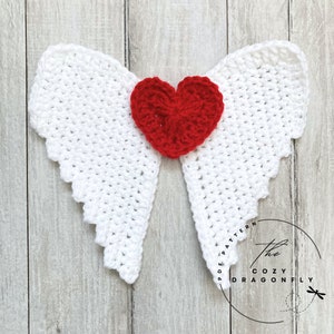 May include: White crocheted angel wings with a red crocheted heart in the center. The wings are on a light wood background. The text "The Cozy Dragonfly" is visible in the lower right corner of the image.