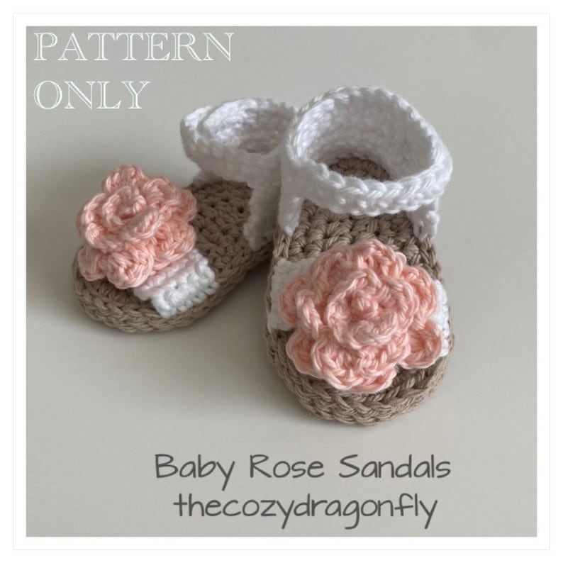 K&ouml;nnte beinhalten: Ein Paar geh&auml;kelte Babysandalen mit einer rosa Blumenverzierung. Die Sandalen sind beige und wei&szlig; mit einem wei&szlig;en Riemen. Der Text "Baby Rose Sandals thecozydragonfly" ist unter den Sandalen gedruckt.
