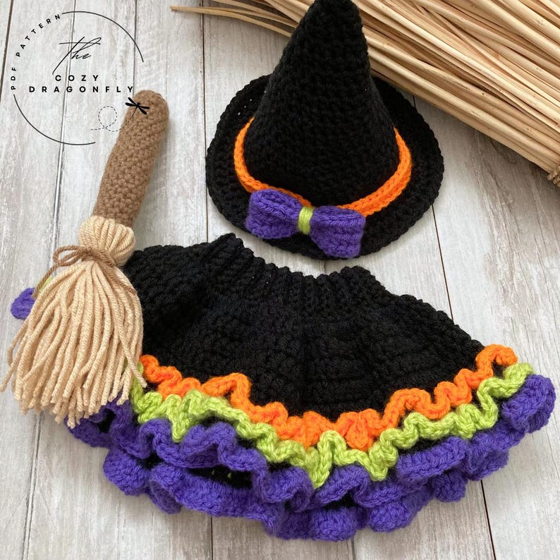 May include: A crochet Halloween costume set featuring a black witch hat with orange trim and a purple bow, a black skirt with orange, green, and purple trim, and a brown and tan broom. The Cozy Dragonfly PDF Pattern logo is in the upper left corner.