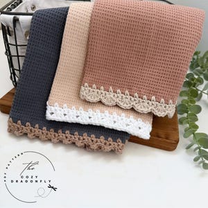 Op de afbeelding: Drie keukenhanddoeken met wafelweefsel in grijs, beige en roze, elk met een decoratieve gehaakte geschulpte rand. De handdoeken zijn netjes op een houten plank gestapeld. Op de achtergrond is een zwarte draadmand te zien.