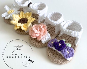May include: Three pairs of crocheted baby sandals with flower embellishments. The sandals are white with brown soles and feature a sunflower, a pink rose, and purple and green flowers.