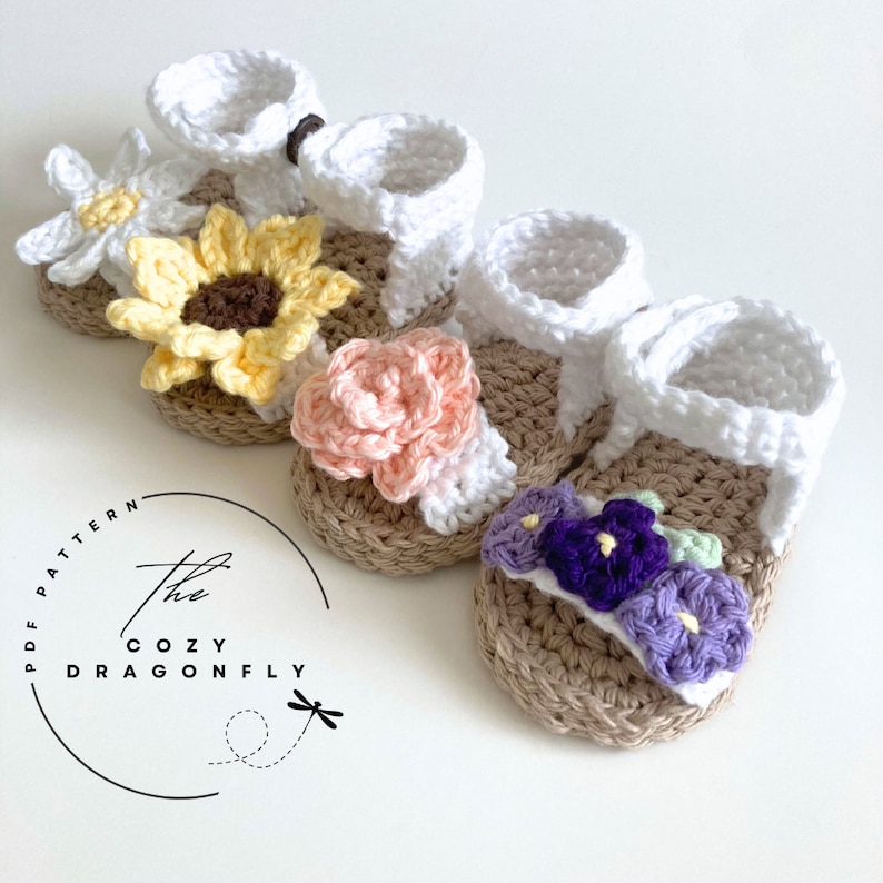 K&ouml;nnte beinhalten: Drei Paar geh&auml;kelte Babysandalen mit Blumenverzierungen. Die Sandalen sind wei&szlig; mit braunen Sohlen und haben eine Sonnenblume, eine rosa Rose und violette und gr&uuml;ne Blumen.