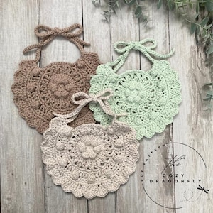 Può includere: Tre bavaglini all'uncinetto in marrone, verde e beige con un bordo a festoni e un motivo floreale al centro. I bavaglini hanno lacci per andare intorno al collo. Il testo "The Cozy Dragonfly PDF Pattern" è visibile nell'angolo in basso a destra dell'immagine.