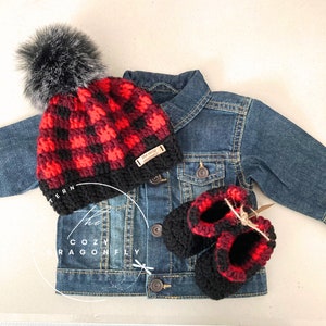 May include: Red and black plaid crochet baby hat with a black faux fur pom pom, matching booties, and a denim jacket. The hat has a label that says "The Cozy Dragonfly".
