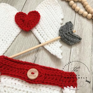 May include: Crochet Cupid costume for a baby, featuring white wings, a red heart, a gray arrow, and a wooden stick. The costume also includes a red crocheted band with a wooden button that says "handmade with love".