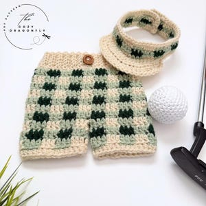 May include: Crocheted golf-themed set in cream, green, and dark green. The set includes shorts, a visor, and a golf ball. The shorts have a button closure. The Cozy Dragonfly logo is in the upper left corner.
