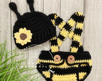 May include: A black crochet hat with two antennae and a yellow sunflower applique, paired with black and yellow striped crochet diaper cover shorts featuring suspenders and two round wooden buttons. Text reads: PDF PATTERN THE COZY DRAGONFLY.