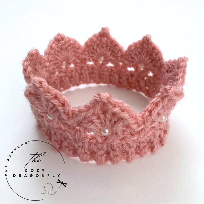 May include: A pink crocheted crown with white pearl beads. The crown is made with a delicate stitch pattern and has a scalloped edge.