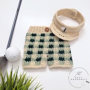 May include: A crocheted golf-themed outfit, including shorts and a visor, in a beige and green checkered pattern. The shorts have a button closure. A golf ball and club are also visible.