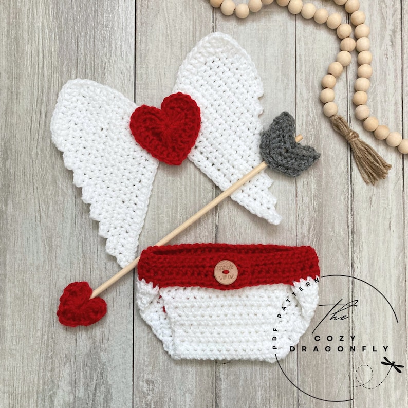 May include: A crochet baby outfit with a red and white diaper, a red heart, and a white crochet arrow with a gray arrowhead. The outfit is on a wooden background with a string of wooden beads.