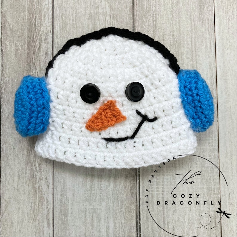 May include: White crocheted snowman hat with blue earmuffs and black button eyes. The hat has a black stitched smile and an orange felt nose.