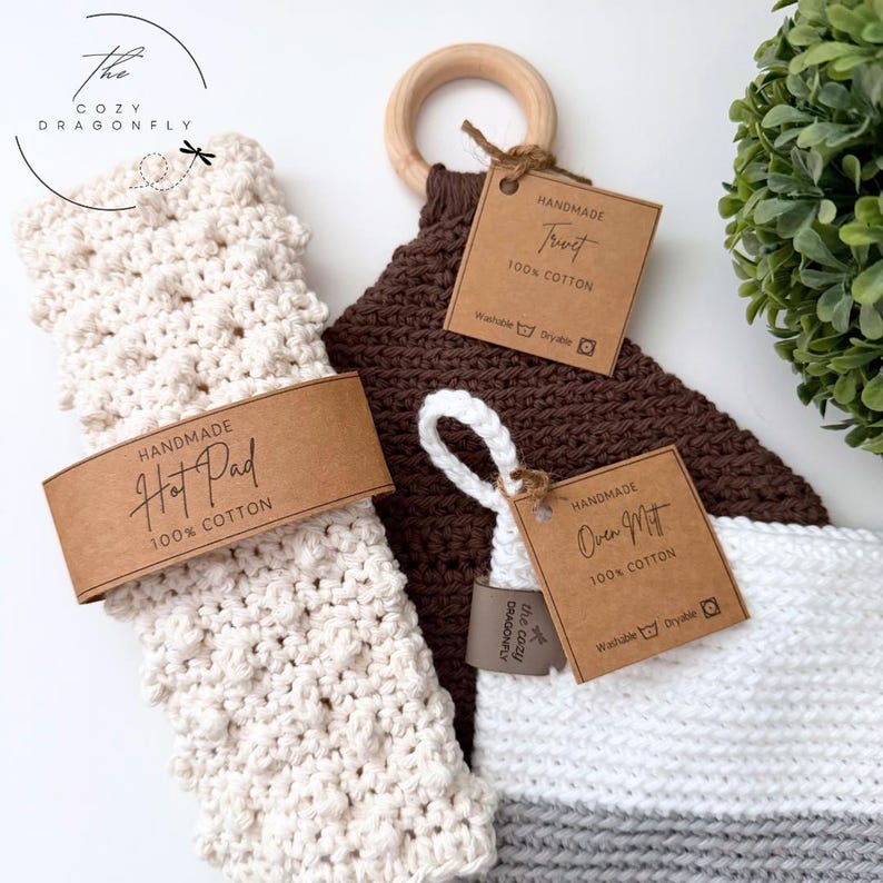 Op de afbeelding: Handgemaakte keukentextiel, waaronder een cr&egrave;mekleurige pannenlap, een bruine onderzetter met een houten ring en een witte en grijze ovenwant. Elk item heeft een bruin label met "Handmade" en "100% Cotton."