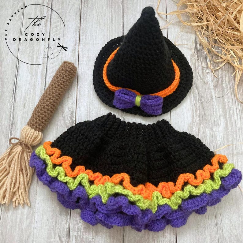May include: A crochet Halloween costume set, including a black witch hat with orange trim and a purple bow, a black skirt with orange, green, and purple trim, and a brown broom. The Cozy Dragonfly pattern is visible in the image.