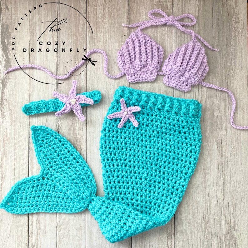 May include: A crocheted mermaid costume for a baby or toddler. The costume includes a purple shell bra top, a turquoise tail with a starfish, and a headband with a starfish.