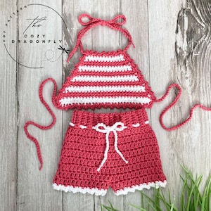 Op de afbeelding: Een gehaakt badpak voor een baby of peuter. Het bovenstuk is halter-stijl met witte strepen op een roze achtergrond. De broek is effen roze met witte afwerking en een witte strik.