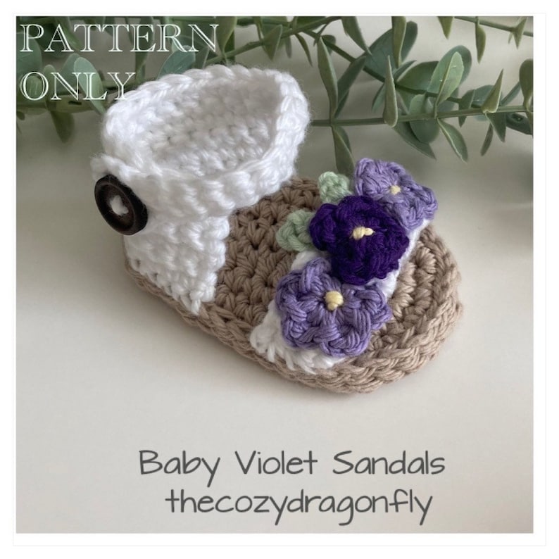 K&ouml;nnte beinhalten: H&auml;kelmuster f&uuml;r Babysandalen mit einem wei&szlig;en Riemen und einer braunen Sohle. Die Sandalen sind mit lila und wei&szlig;en Blumen verziert.