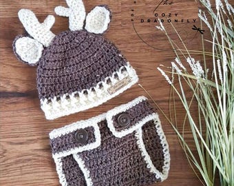 May include: A dark brown crochet hat with cream-colored antlers and ears, paired with matching dark brown crochet diaper cover shorts featuring cream trim and two dark brown buttons. The hat includes a small rectangular tag with text.