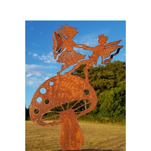 May include: Two fairies dancing on top of a rusty metal mushroom sculpture. The fairies are holding hands and have wings. The mushroom is brown and has white spots.