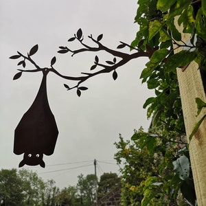 May include: A black metal silhouette of a bat hanging upside down from a tree branch. The silhouette is attached to a wooden post.