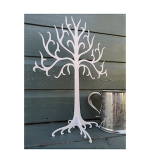 May include: White wooden tree-shaped jewellery stand with intricate branches and a base with swirling roots. The stand is on a green wooden surface.