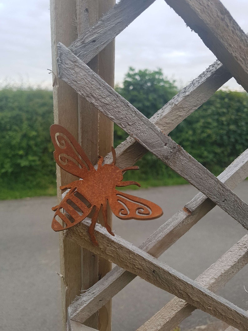 K&ouml;nnte beinhalten: Eine rostige Metallbienen-Skulptur, die an einem Holzgitter befestigt ist. Die Biene ist mit Fl&uuml;geln und einem gestreiften K&ouml;rper detailliert.