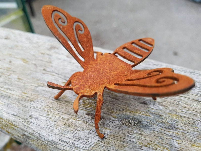 K&ouml;nnte beinhalten: Eine rostige Metall-Bienenskulptur mit filigranen Details an den Fl&uuml;geln. Die Biene befindet sich in Flugposition mit ausgebreiteten Fl&uuml;geln.