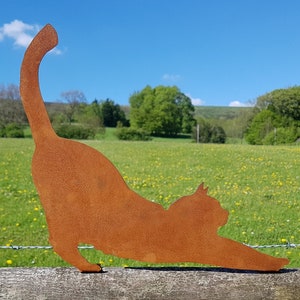 May include: A rusty metal silhouette of a cat stretching in a yoga pose. The silhouette is placed on a wooden fence post in front of a green field with yellow wildflowers.