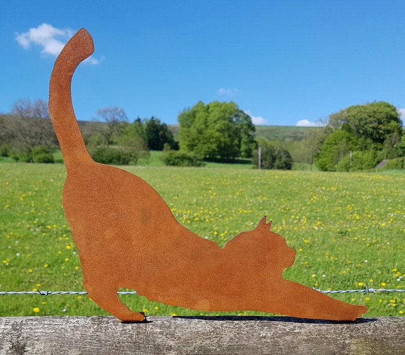 May include: A rusty metal silhouette of a cat stretching in a yoga pose. The silhouette is placed on a wooden fence post in front of a green field with yellow wildflowers.