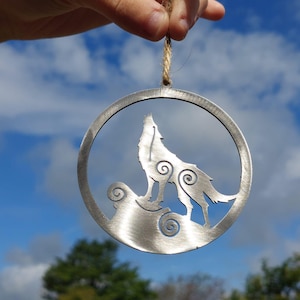 May include: A circular metal ornament depicting a howling wolf silhouette. The wolf is positioned on a stylized rock, with decorative swirls. The ornament hangs from a natural twine cord against a blue sky with clouds.