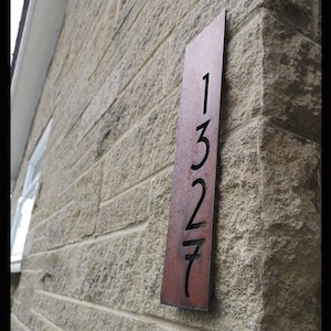 May include: A rectangular, reddish-brown metal address sign displaying the numbers "1327" cut out. The sign is mounted on a light-coloured stone wall. The numbers are clearly visible, offering a modern aesthetic.